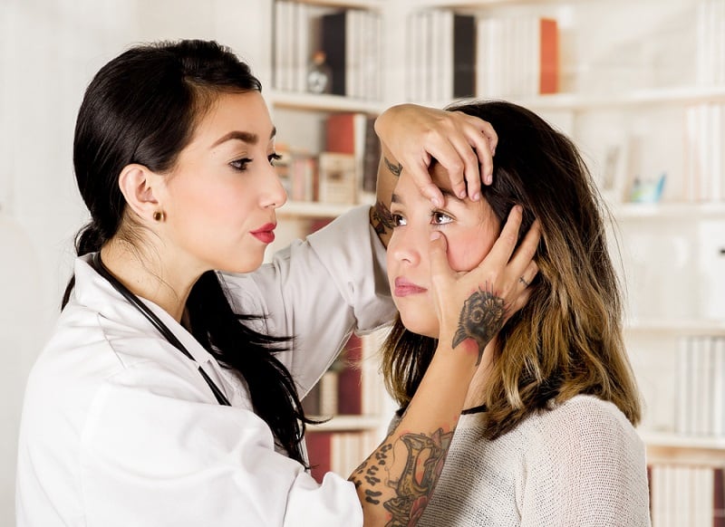 Hermosa joven doctora tatuada usando su mano para revisar los ojos de su paciente